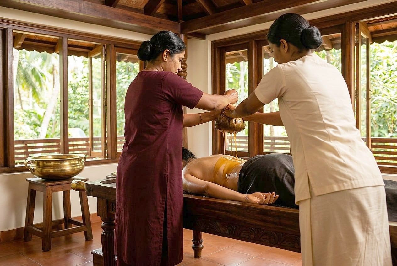 Two therapists squeeze warm oil from cloths over a person's back on a wooden table