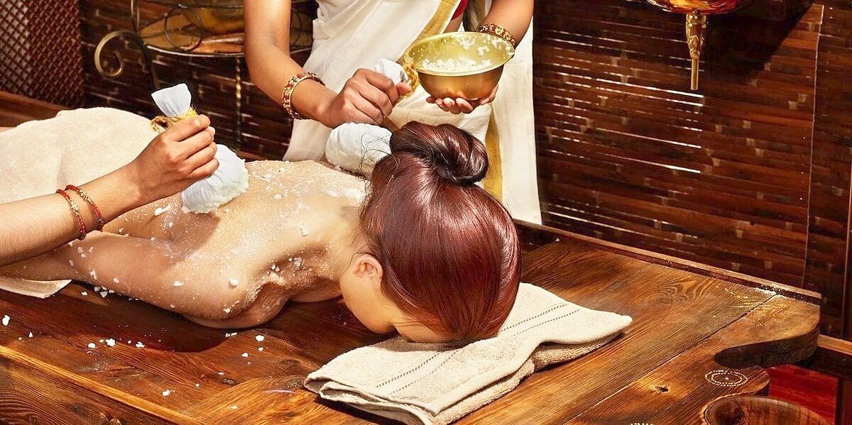 Two therapists warming the back with herbal boluses, a brass bowl in hand