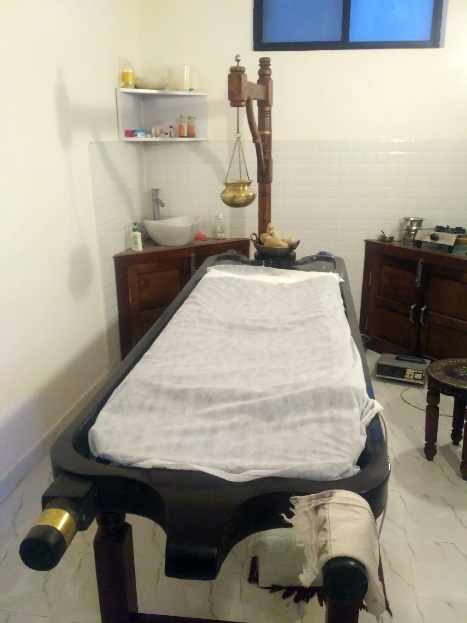 A treatment table with a brass shirodhara vessel and a shelf of oils